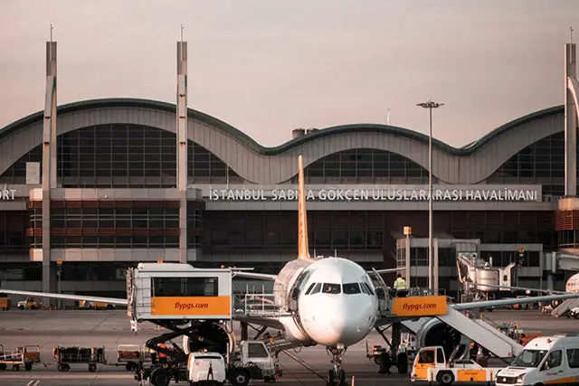 A commercial airliner is parked at a gate with ground support vehicles around it, in front of an airport with the text "ISTANBUL SABIHA GOKCEN ULUSLARARASI HAVALIMANI".
