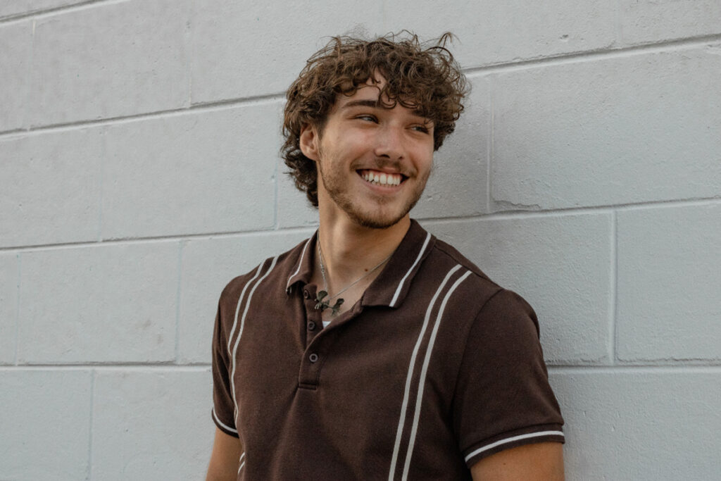young man with curly hair smiling while leaning against a white brick wall